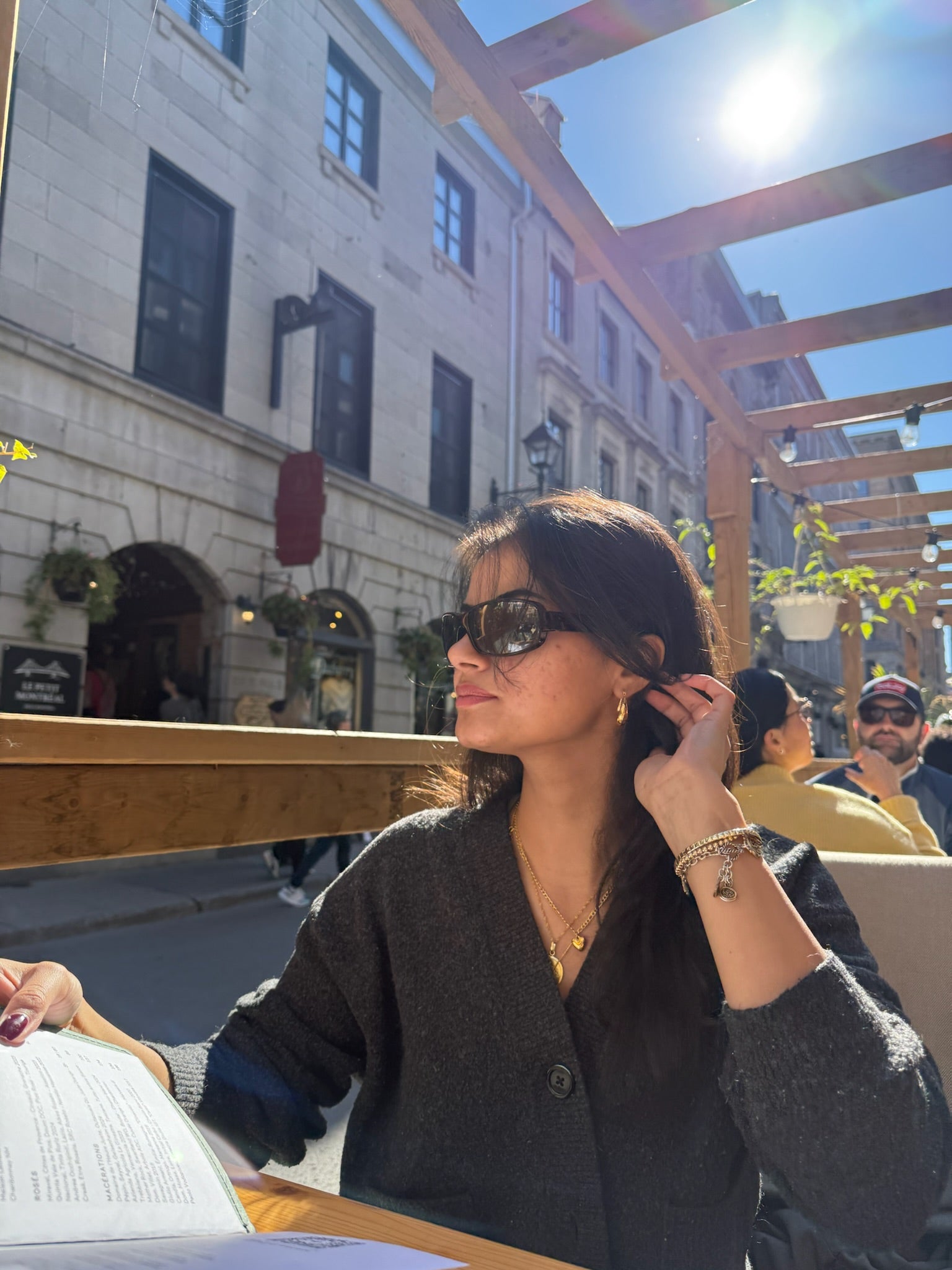 Woman sitting outdoors in an urban setting, wearing sunglasses, a stack of gold necklaces, a pair of dainty everyday gold earrings  and a dark coat 